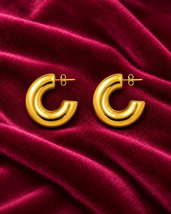 Elli Jewellery chunky C-shaped hoop earrings in gold plated metal resting on a luxurious deep red velvet fabric.