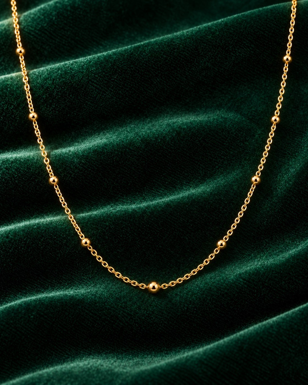 Elli Jewellery gold plated dainty ball bead chain necklace on a dark green velvet background