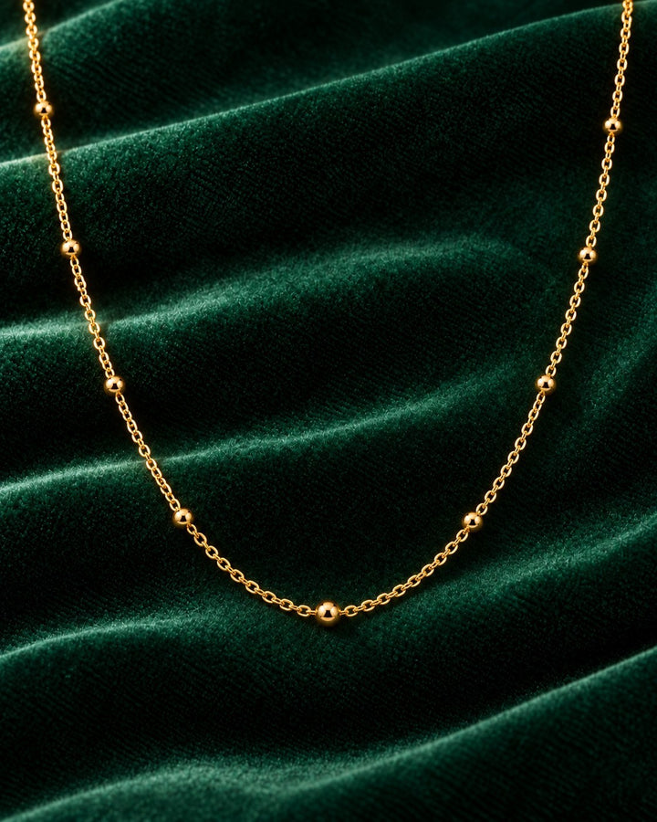 Elli Jewellery gold plated dainty ball bead chain necklace on a dark green velvet background