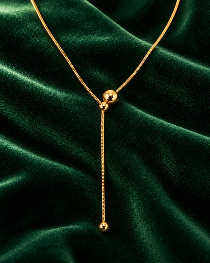 Elli Jewellery gold plated minimalist lariat necklace with hanging ball pendants on a dark green velvet background