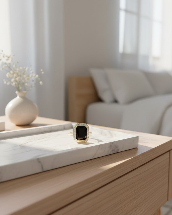 Elli Jewellery gold plated chunky ring with a black rectangular stone on a marble tray in a bright bedroom setting.