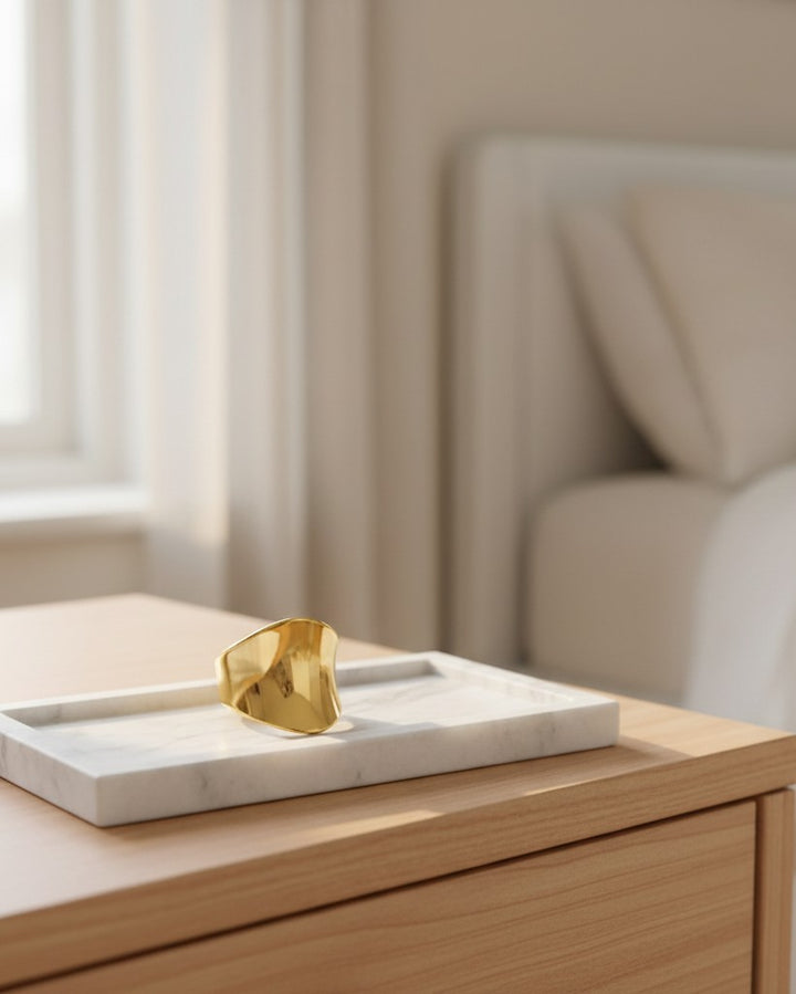 Elli Jewellery gold plated brushed cambered open cuff ring resting on a white marble tray in a minimalist bedroom.