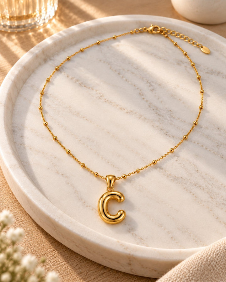 Elli Jewellery gold plated necklace featuring a chunky letter C pendant on a delicate beaded chain on a marble tray.