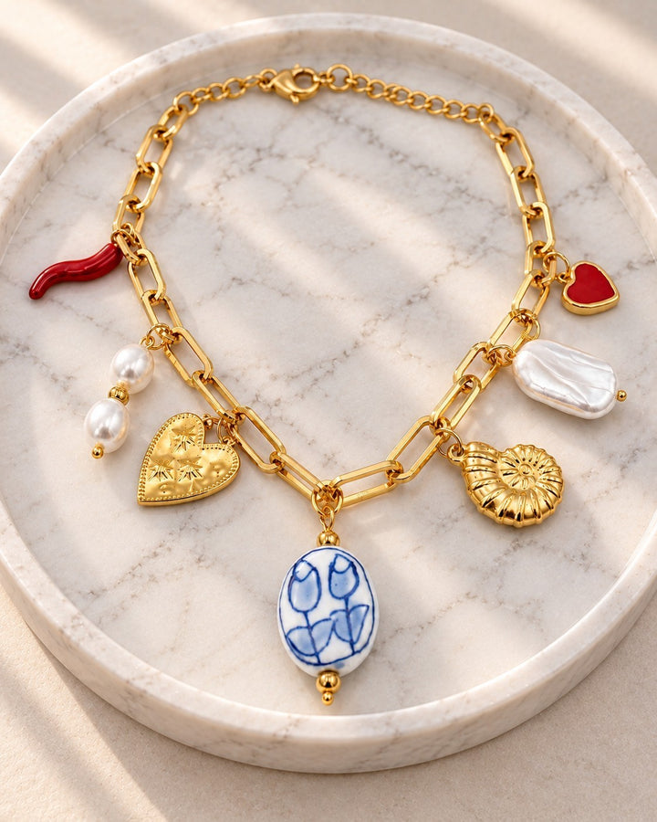 Elli Jewellery gold plated charm bracelet featuring a shell, heart, pearl, and blue floral ceramic pendant on a tray.