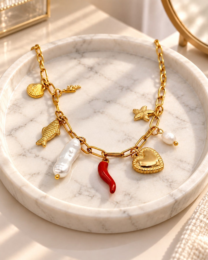 Elli Jewellery gold-plated charm necklace featuring a heart, pearl, red chili, and seashell charms on a marble tray.