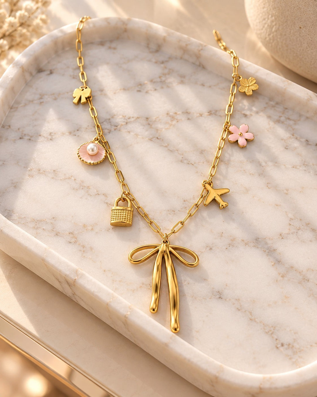 Elli Jewellery gold plated charm necklace featuring a ribbon bow, padlock, airplane, and floral accents on a tray.