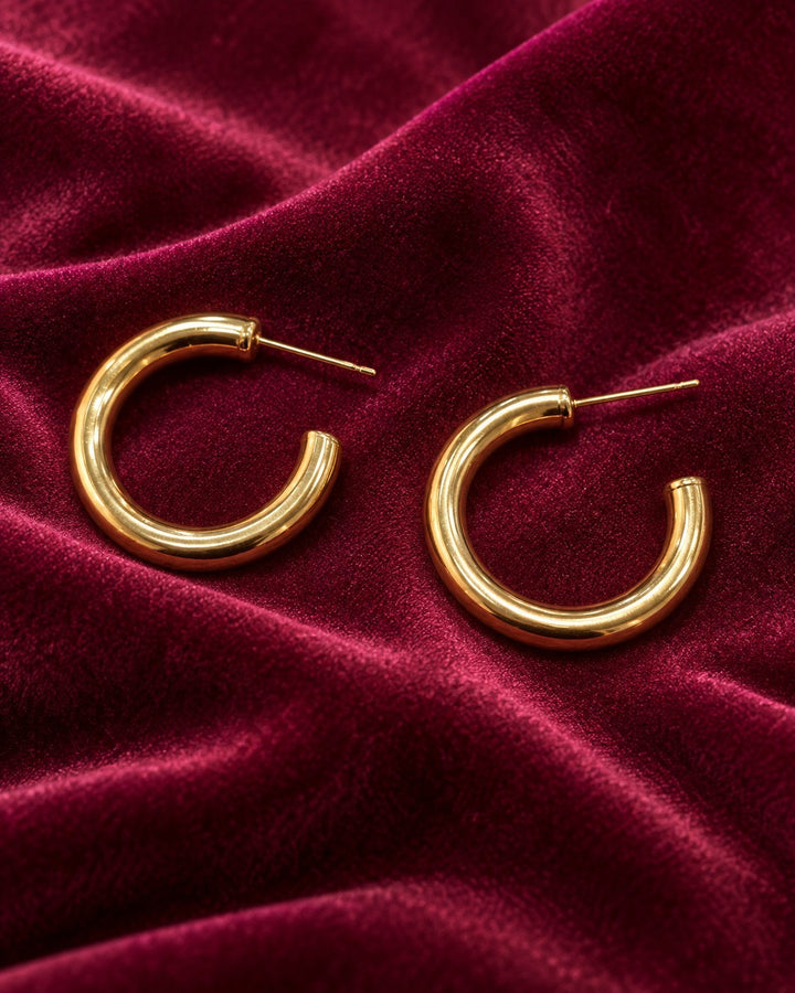 Elli Jewellery gold plated chunky hoop earrings with a polished finish resting on a deep red velvet fabric background.