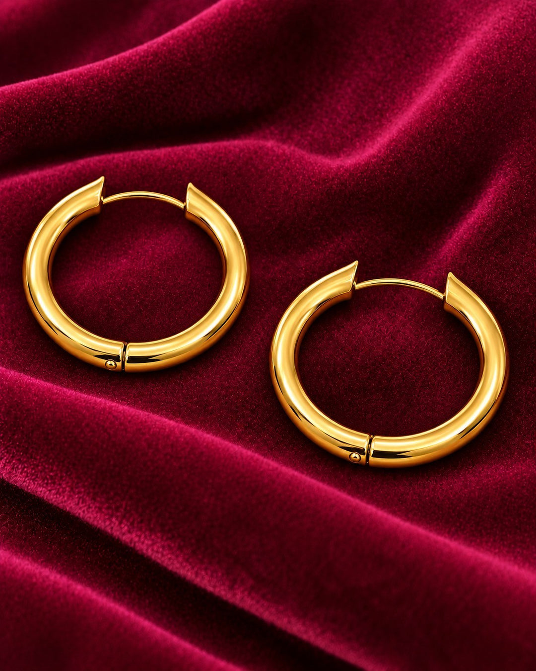 Elli Jewellery gold plated chunky huggie hoop earrings resting on a luxurious deep red velvet fabric background.