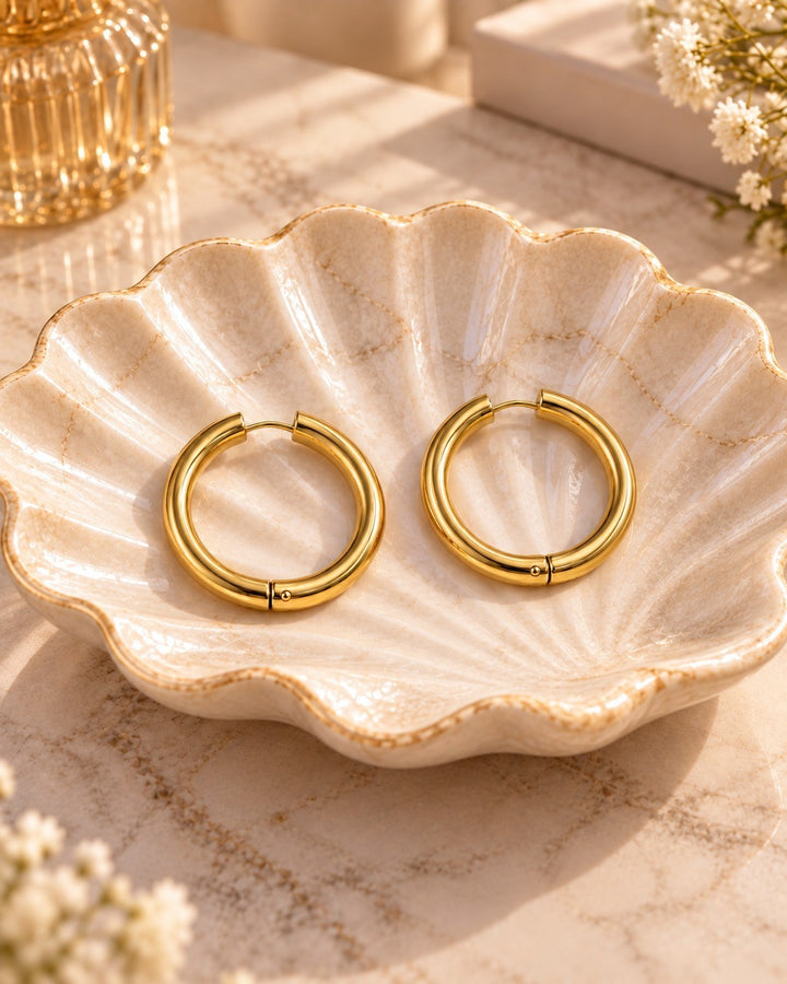 Elli Jewellery gold plated chunky huggie hoop earrings resting in a decorative ceramic shell dish.