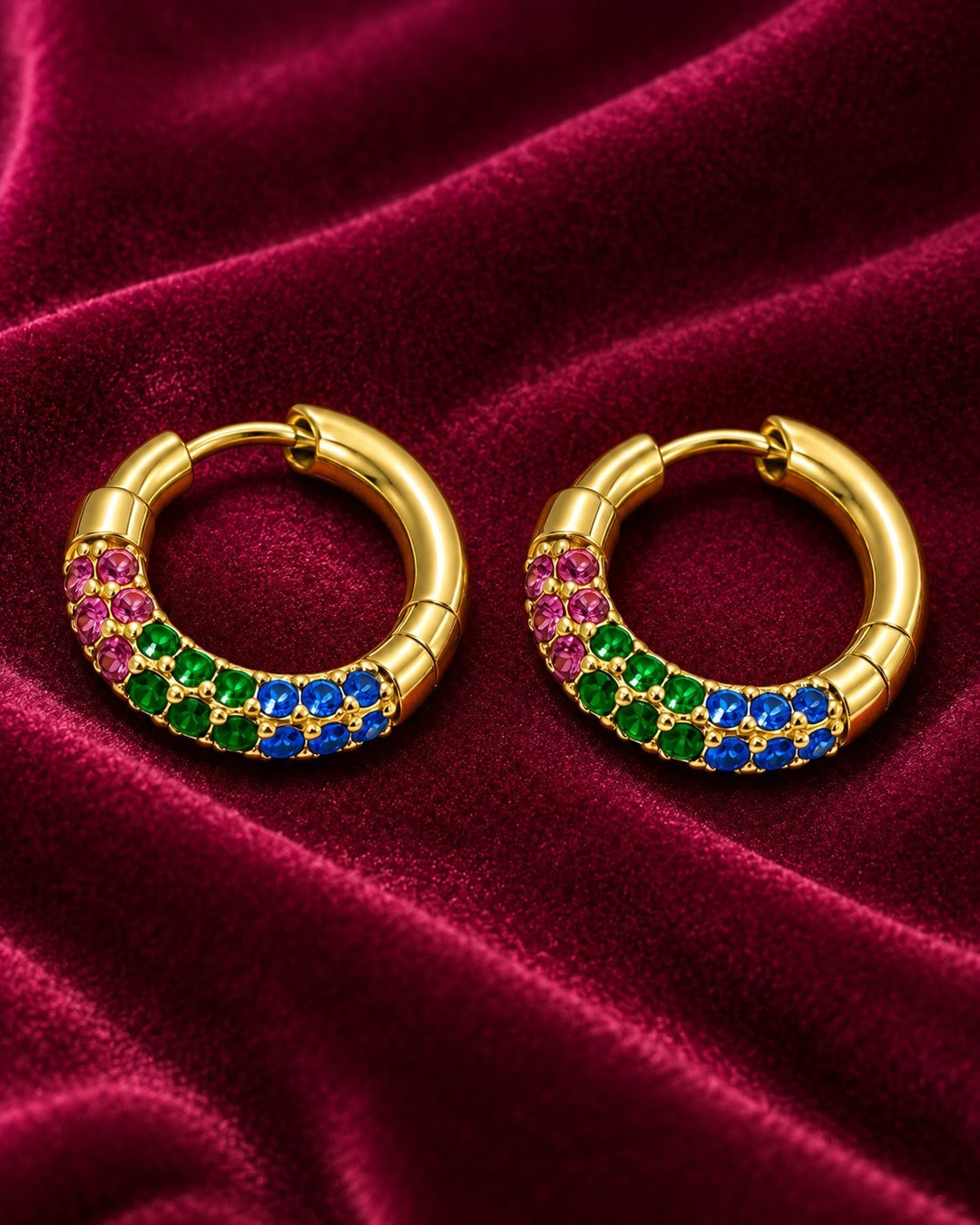 Elli Jewellery gold plated hoop earrings featuring vibrant pink, green, and blue crystals on a deep red velvet fabric.