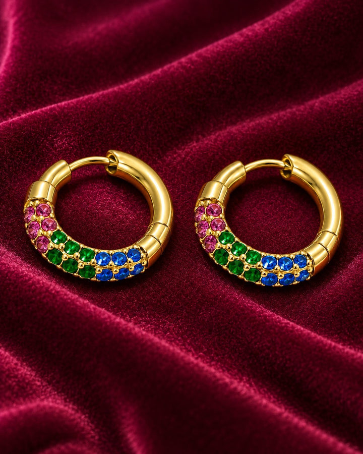 Elli Jewellery gold plated hoop earrings featuring vibrant pink, green, and blue crystals on a deep red velvet fabric.
