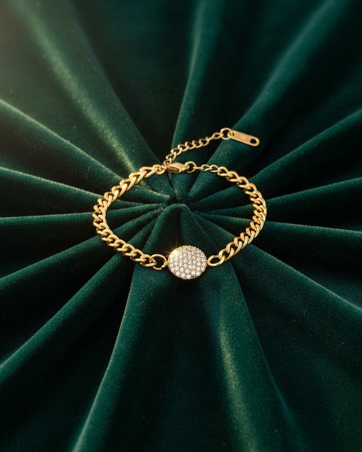 Elli Jewellery gold plated curb chain bracelet featuring a round diamanté pendant on a dark green velvet background.