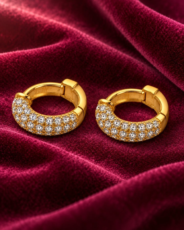 Elli Jewellery gold plated huggie hoop earrings featuring sparkling diamanté stones on a deep red velvet background.