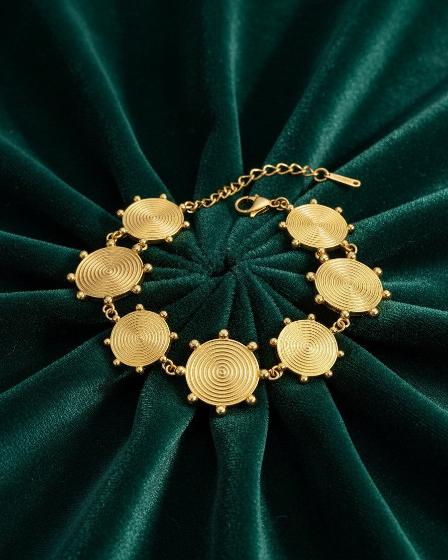 Elli Jewellery gold plated charm bracelet with circular textured discs on a dark green velvet background