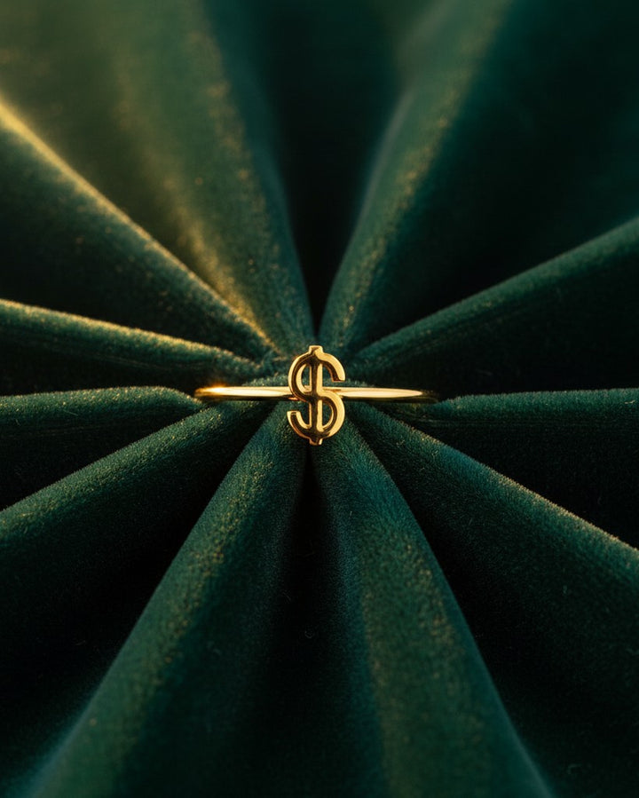 Elli Jewellery gold plated waterproof statement ring featuring a dollar sign symbol on a dark green velvet background.