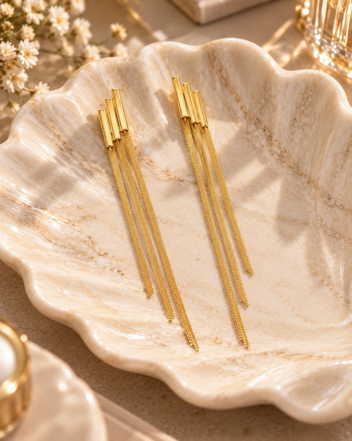 Elli Jewellery gold plated long drop chain earrings resting on a decorative shell dish in warm sunlight.