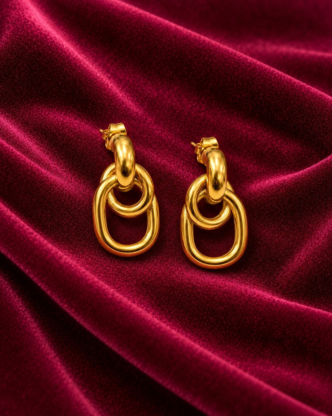 Elli Jewellery gold plated waterproof drop earrings with interlocking oval links on a deep red velvet background.
