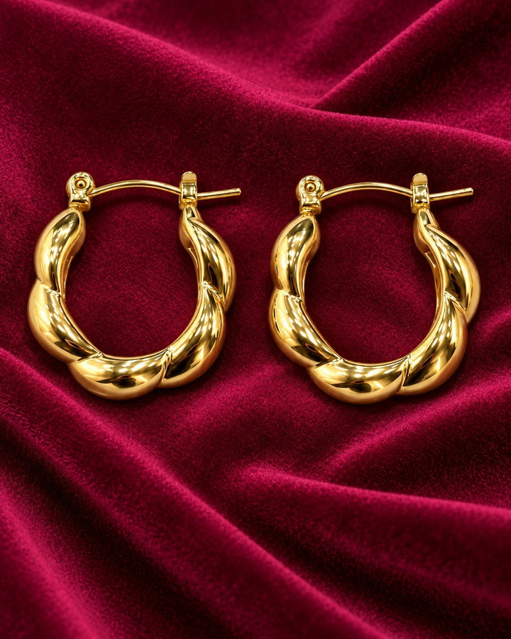 Elli Jewellery gold plated chunky geometric hoop earrings with a twisted sculptural design on deep red velvet fabric.