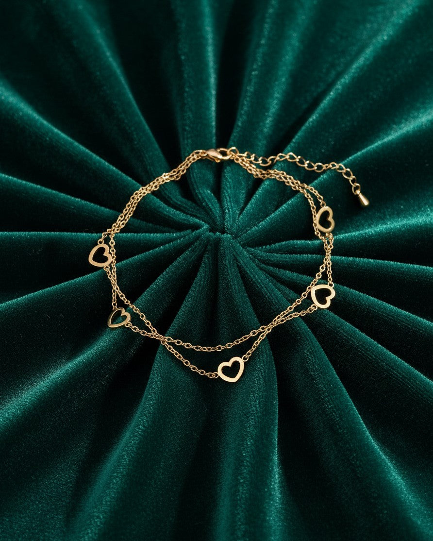 Elli Jewellery gold-toned double chain anklet featuring delicate heart charms on a dark green velvet background.
