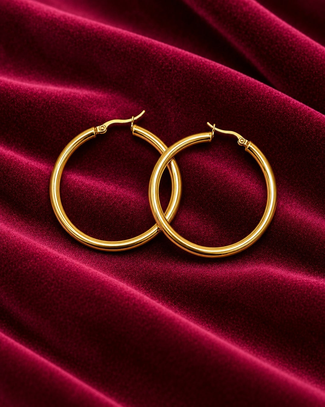 Elli Jewellery 40mm gold-toned hoop earrings resting on a luxurious deep red velvet fabric background.
