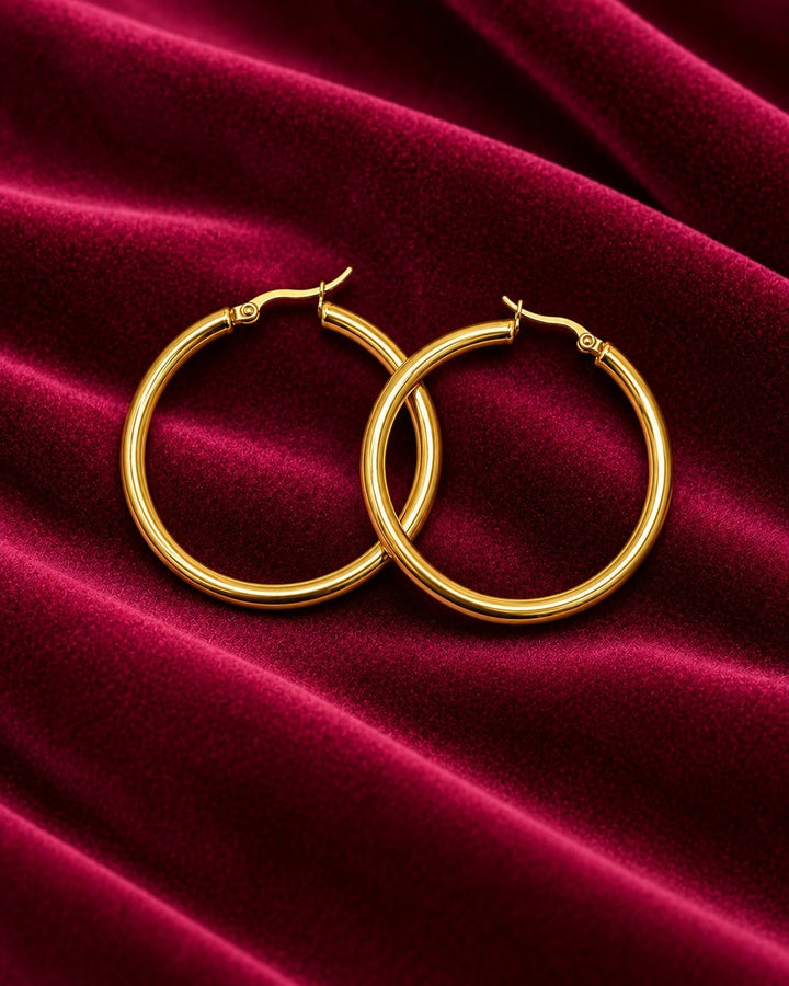 Elli Jewellery 40mm gold-toned hoop earrings resting on a luxurious deep red velvet fabric background.