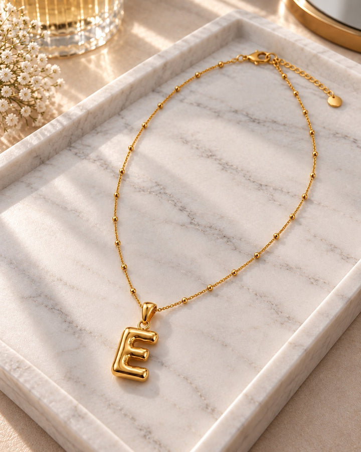 Elli Jewellery gold plated waterproof chunky initial E pendant necklace on a marble tray with soft sunlight.