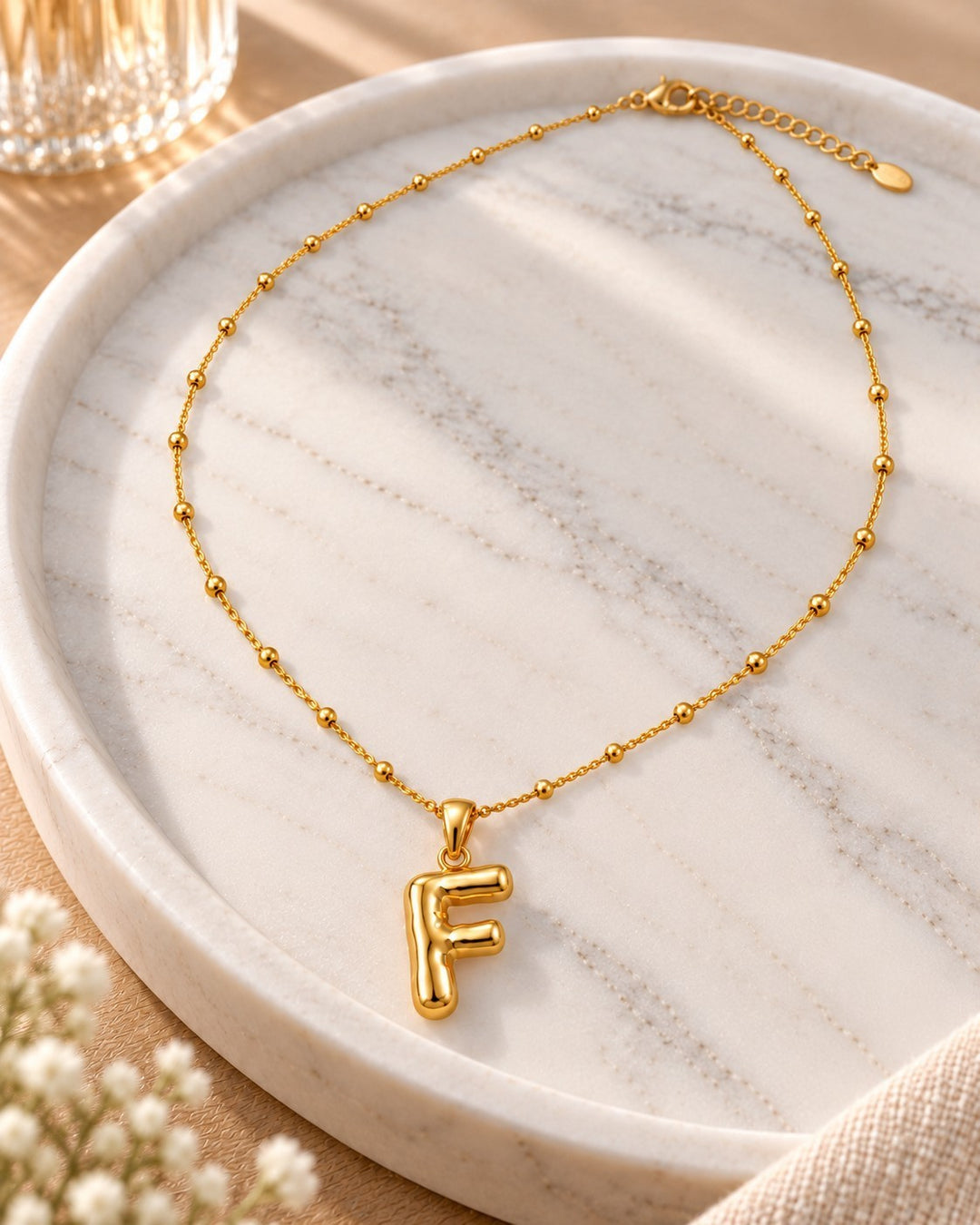 Elli Jewellery gold plated chunky initial F necklace with a delicate beaded chain on a marble tray.