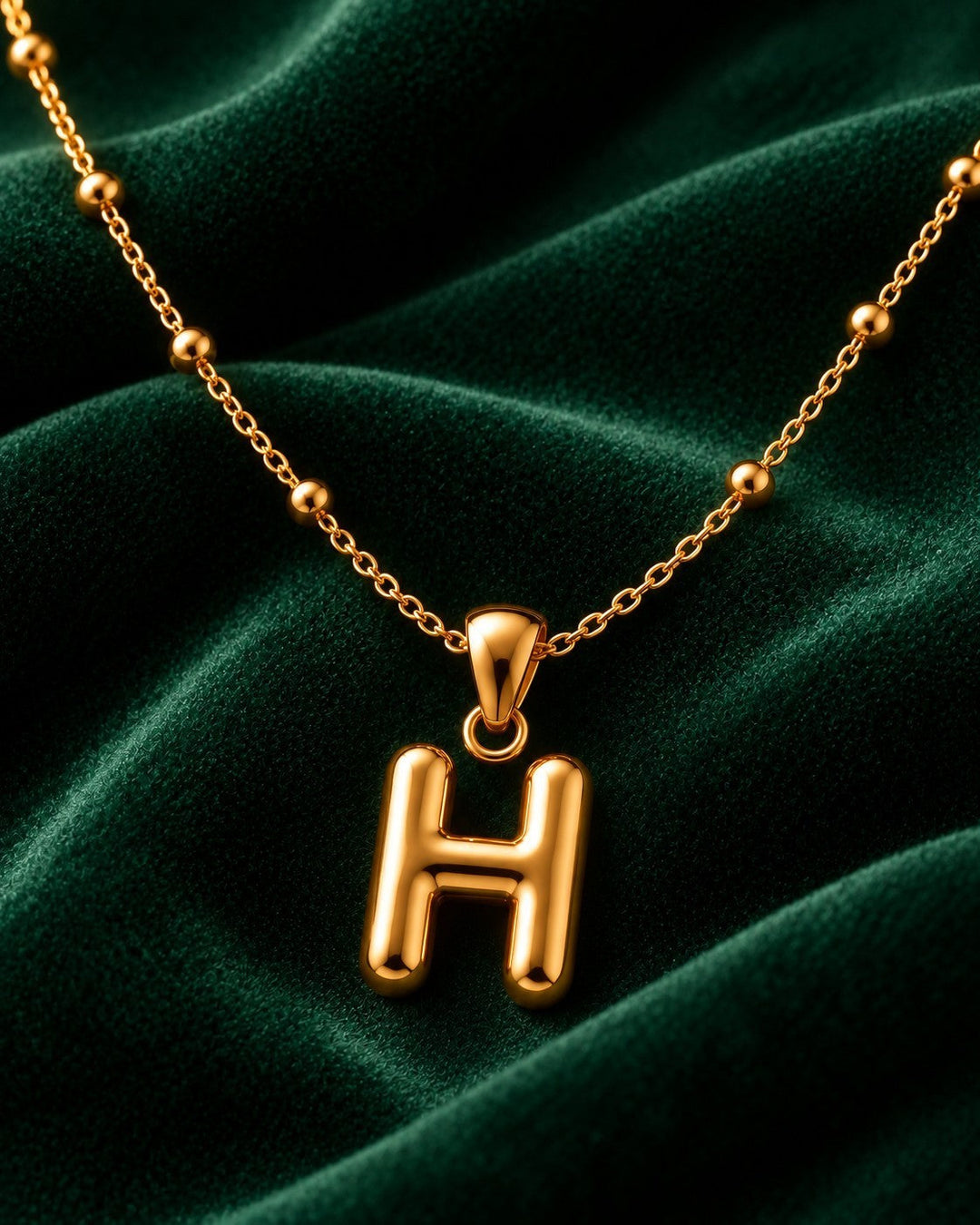 Elli Jewellery gold plated chunky initial H pendant necklace on a delicate beaded chain against dark green velvet.