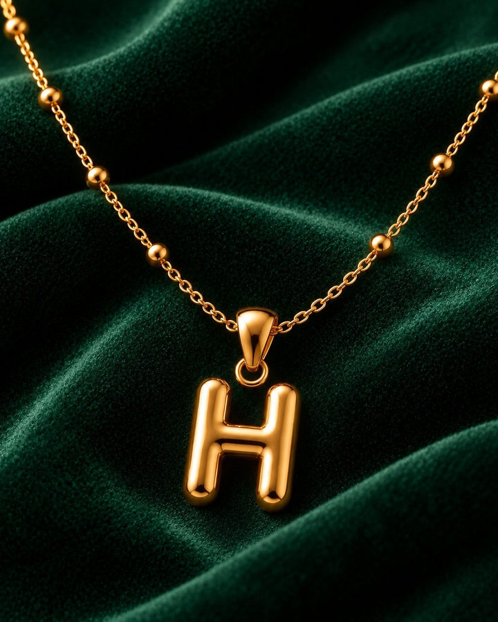 Elli Jewellery gold plated chunky initial H pendant necklace on a delicate beaded chain against dark green velvet.