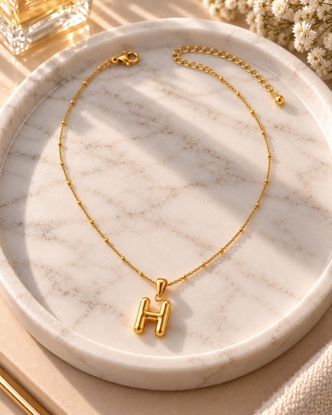 Elli Jewellery gold plated chunky initial H pendant necklace on a delicate beaded chain displayed on a marble tray.