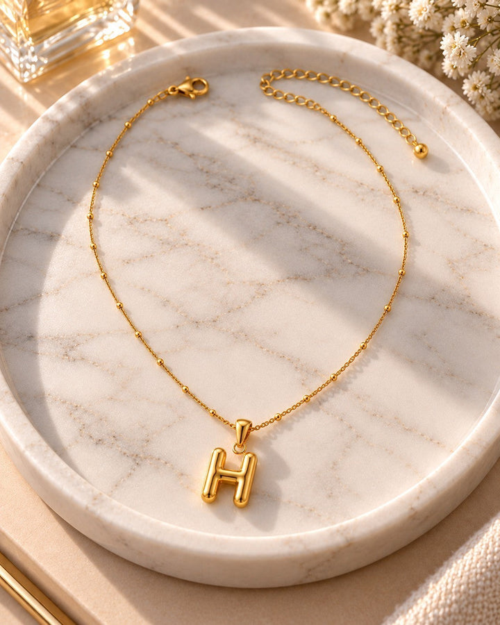 Elli Jewellery gold plated chunky initial H pendant necklace on a delicate beaded chain displayed on a marble tray.