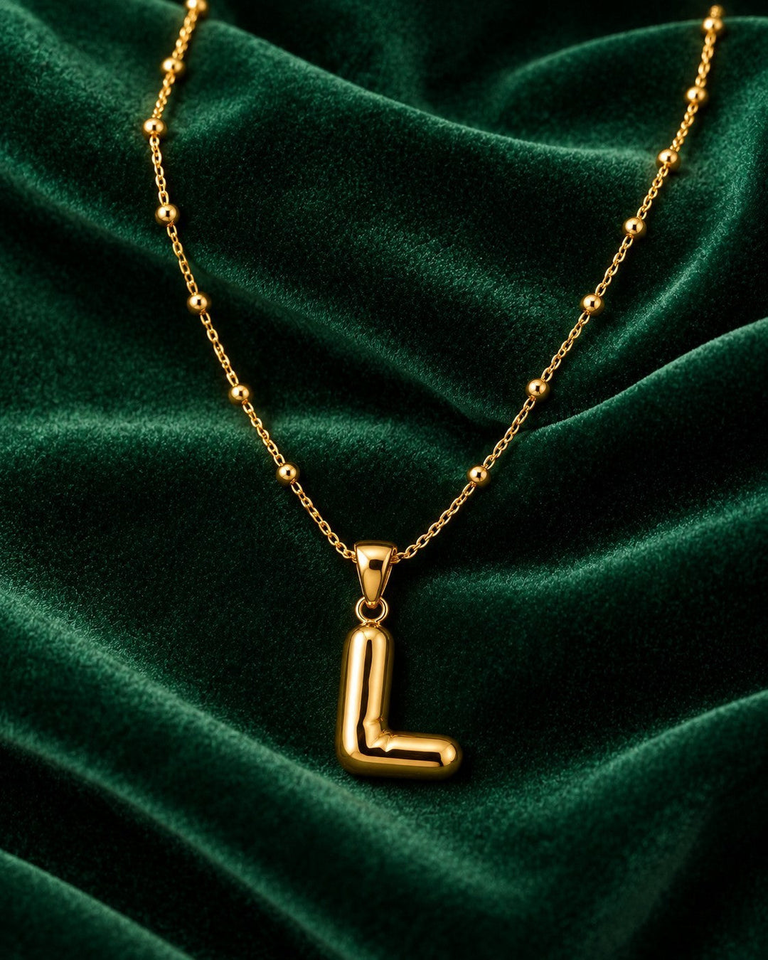 Elli Jewellery gold plated initial L pendant necklace on a delicate beaded chain against a dark green velvet background