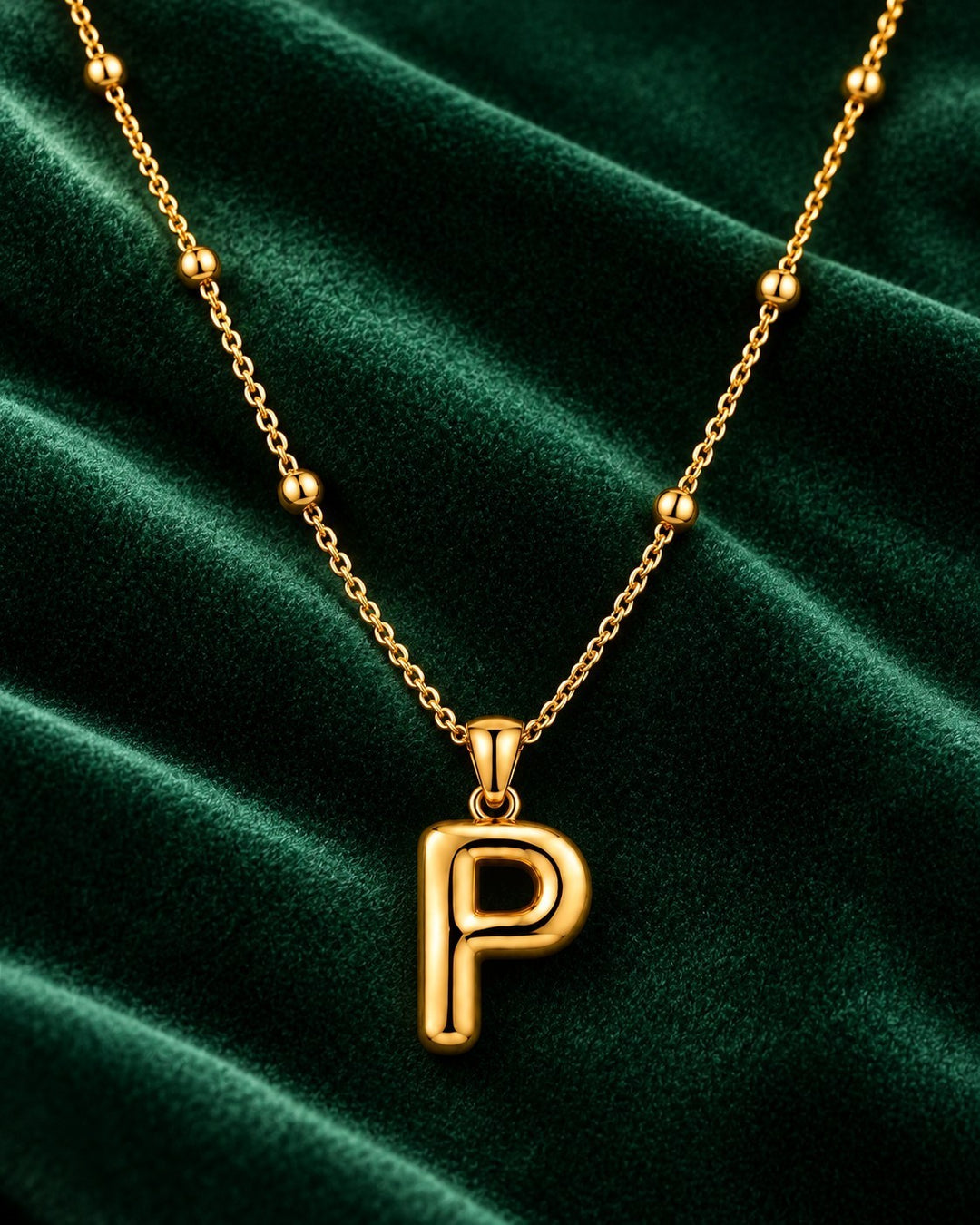 Elli Jewellery gold plated chunky initial P pendant necklace on a beaded chain against a dark green velvet background