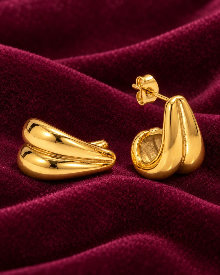 Elli Jewellery gold plated stainless steel interlocking double C hoop earrings resting on deep red velvet fabric.