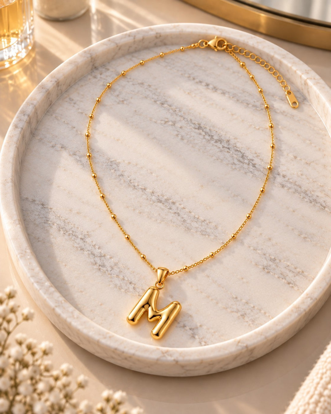 Elli Jewellery gold toned letter M pendant necklace on a delicate beaded chain resting on a marble tray.