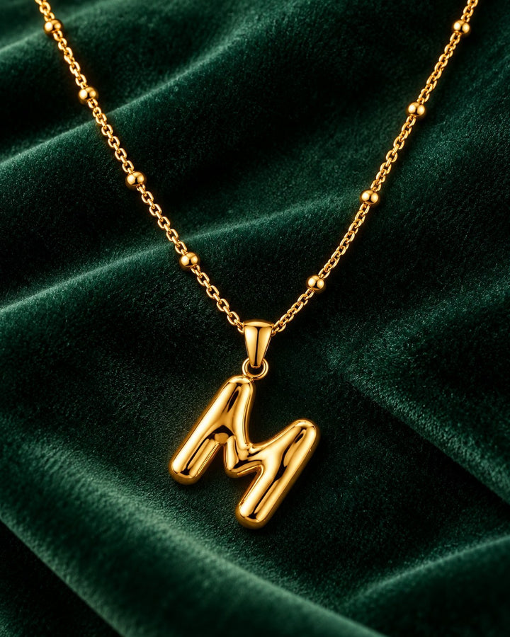 Elli Jewellery gold toned letter M pendant necklace on a beaded chain against a dark green velvet background