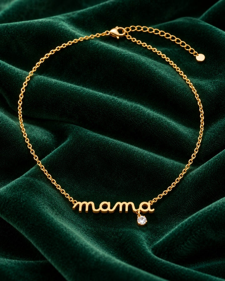 Elli Jewellery gold plated dainty MAMA script pendant necklace with a small crystal accent on green velvet fabric.
