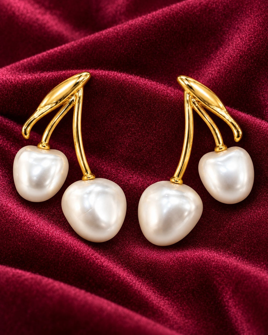 Elli Jewellery gold-toned cherry-shaped drop earrings with lustrous white pearls on a deep red velvet background.