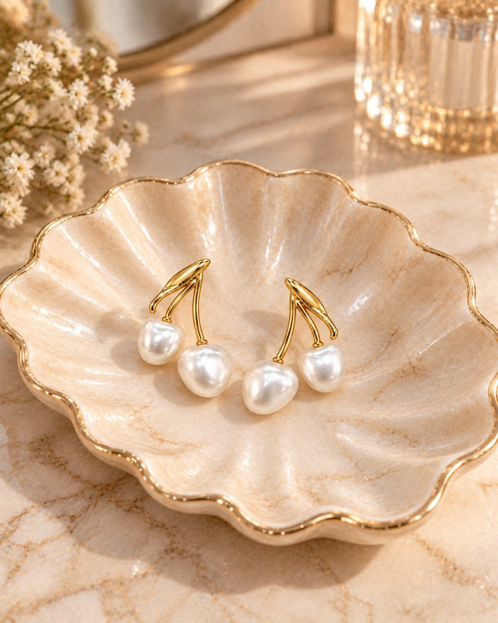Elli Jewellery gold toned cherry drop earrings with lustrous pearl-effect beads resting in a decorative shell dish.