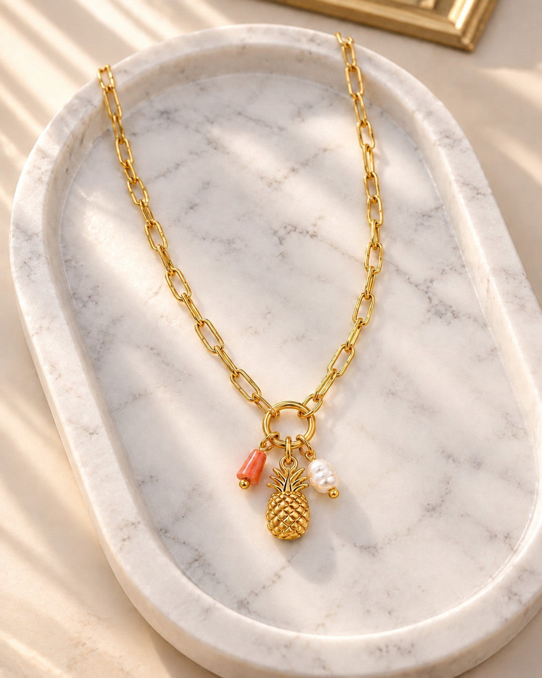 Elli Jewellery gold plated pineapple pendant necklace with a pearl and coral charm on a marble tray.