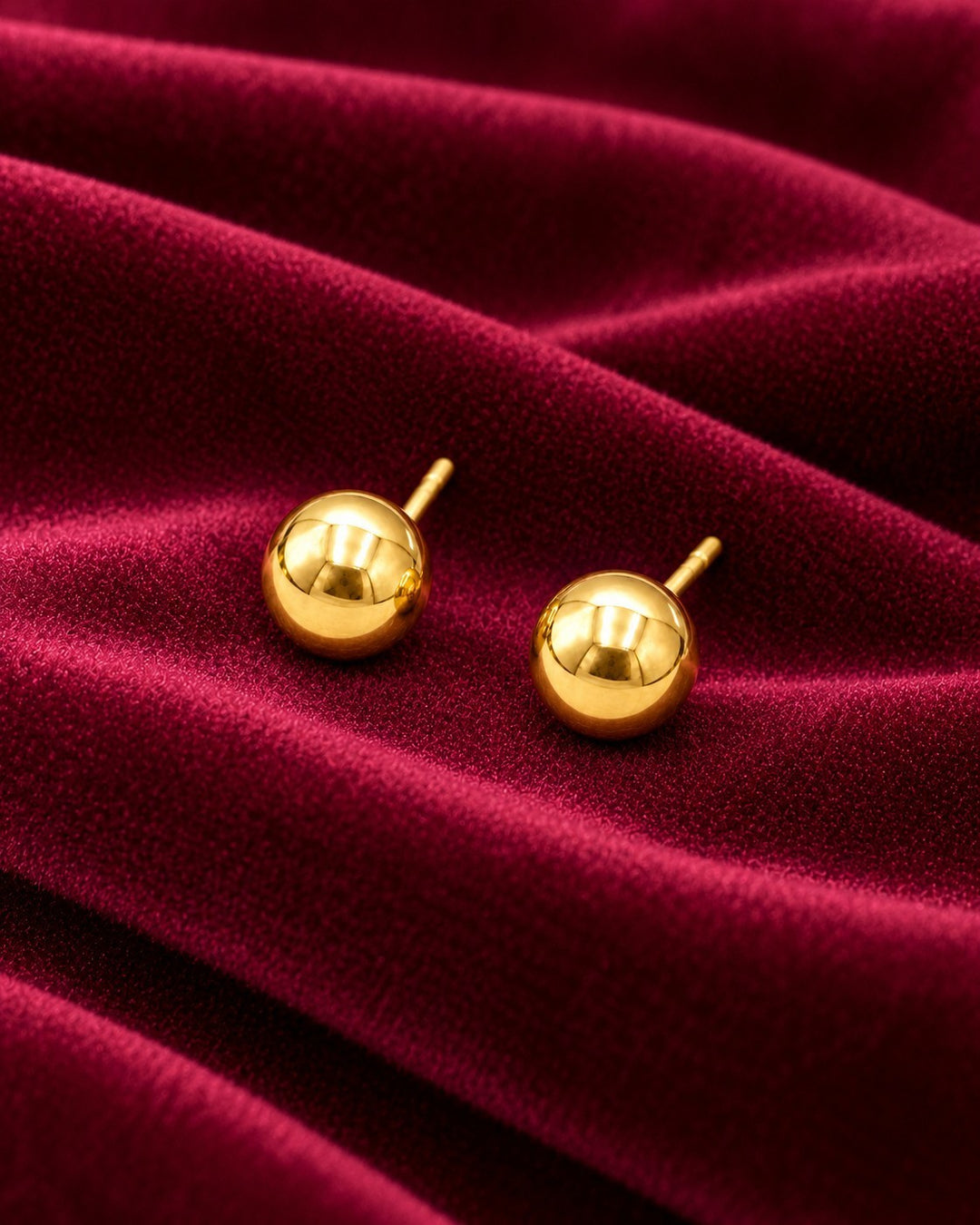 Elli Jewellery 6mm round gold-plated stud earrings resting on a luxurious deep red velvet fabric background.