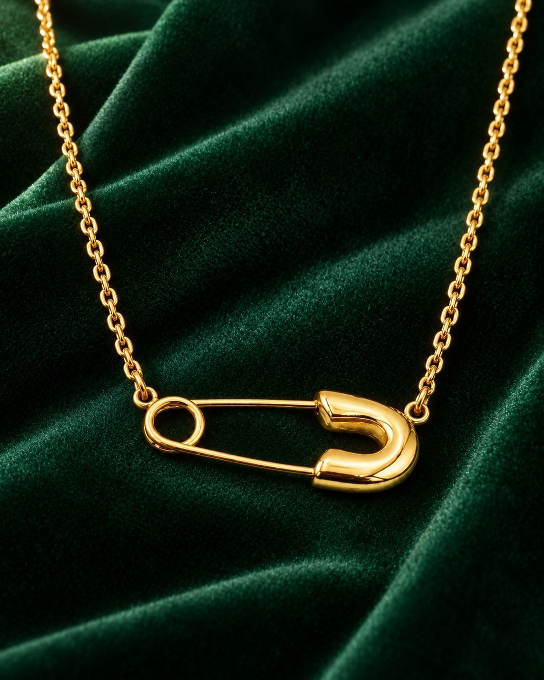 Elli Jewellery gold plated safety pin pendant on a dainty chain resting on a dark green velvet background