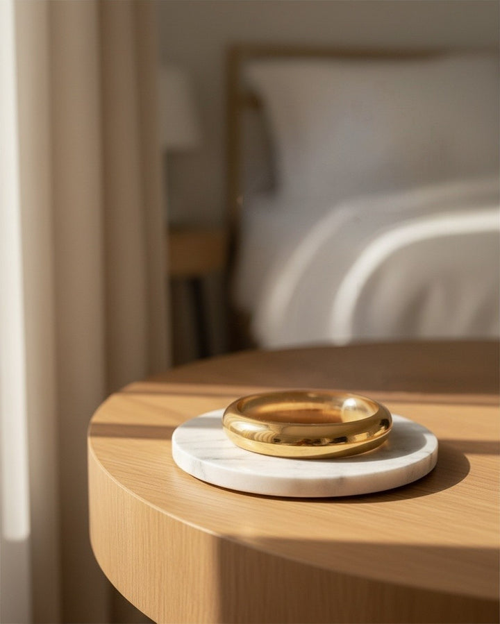 Elli Jewellery gold plated 2cm slim bangle resting on a white marble coaster on a wooden table in soft sunlight.