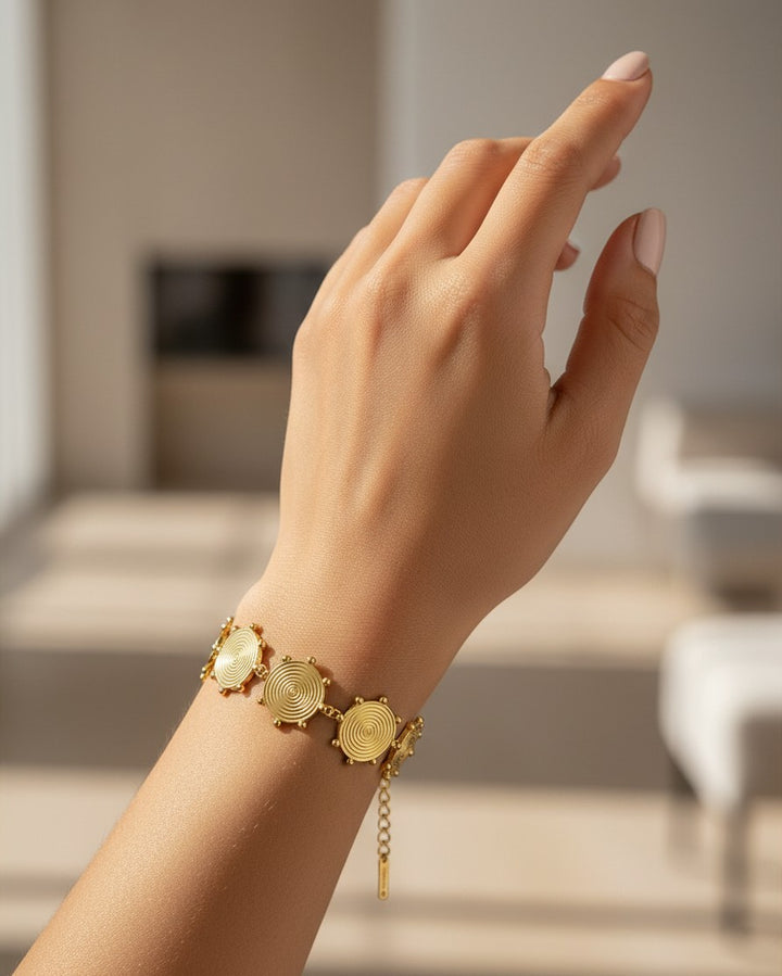Elli Jewellery gold plated charm bracelet with circular spiral motifs worn on a woman's wrist in a bright room