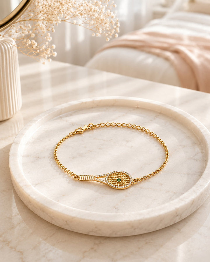 Elli Jewellery gold plated tennis racquet charm bracelet with pavé crystals and a green crystal ball on a marble tray.