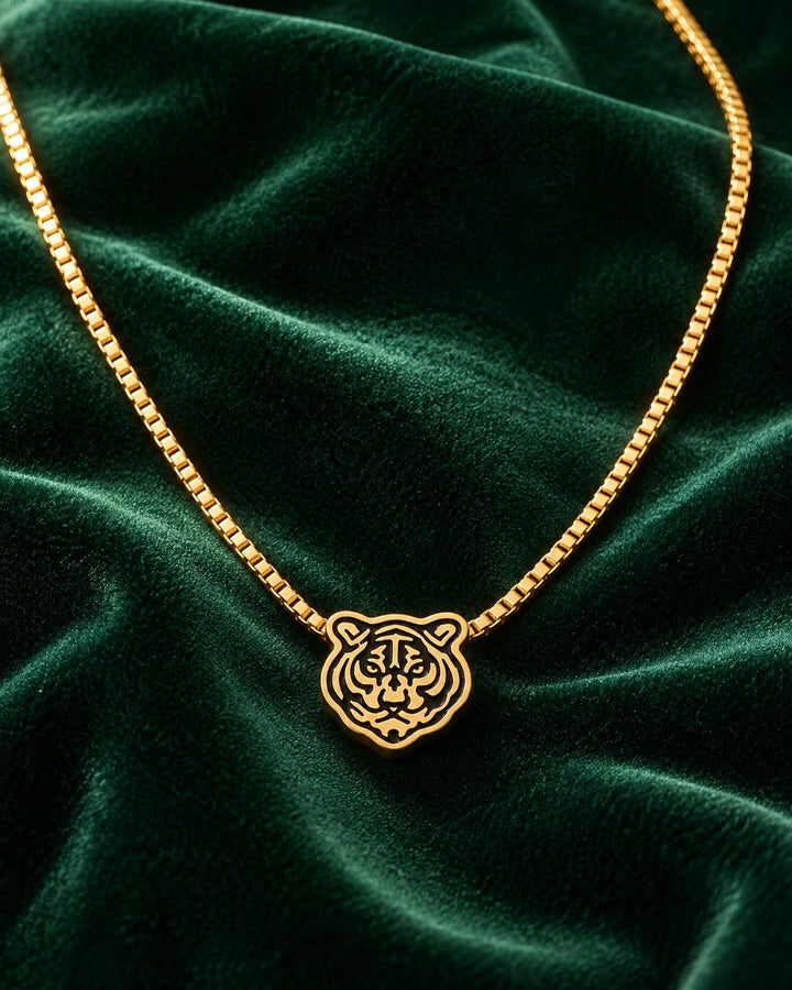 Elli Jewellery gold plated tiger face pendant on a delicate box chain resting on a dark green velvet background.