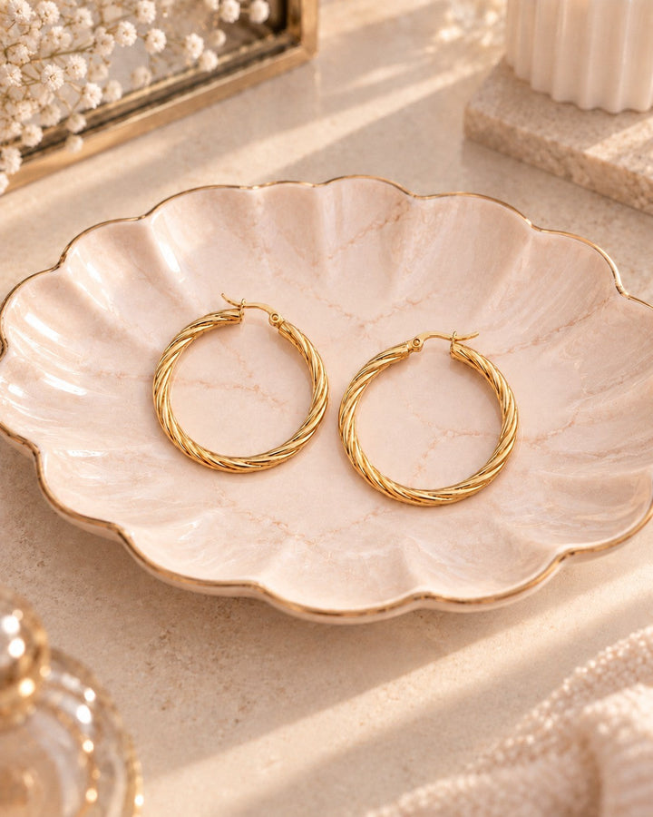 Elli Jewellery large gold-toned hoop earrings with a twisted rope texture resting on a decorative scalloped dish.