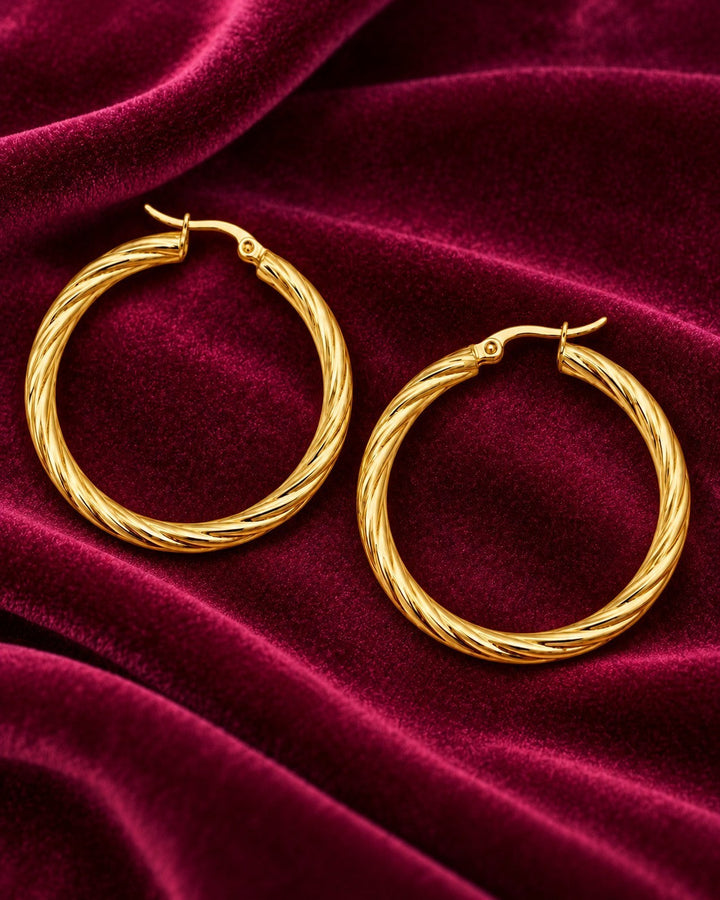 Elli Jewellery gold-toned large hoop earrings with a twisted texture, displayed on a luxurious deep red velvet fabric.