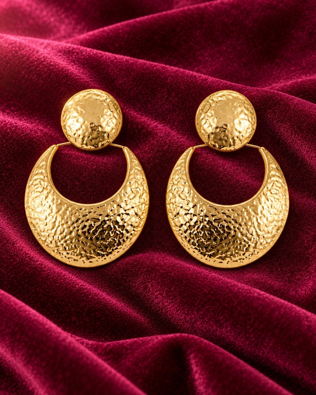 Elli Jewellery hammered crescent drop earrings in 18ct gold plated metal displayed on a rich red velvet background.