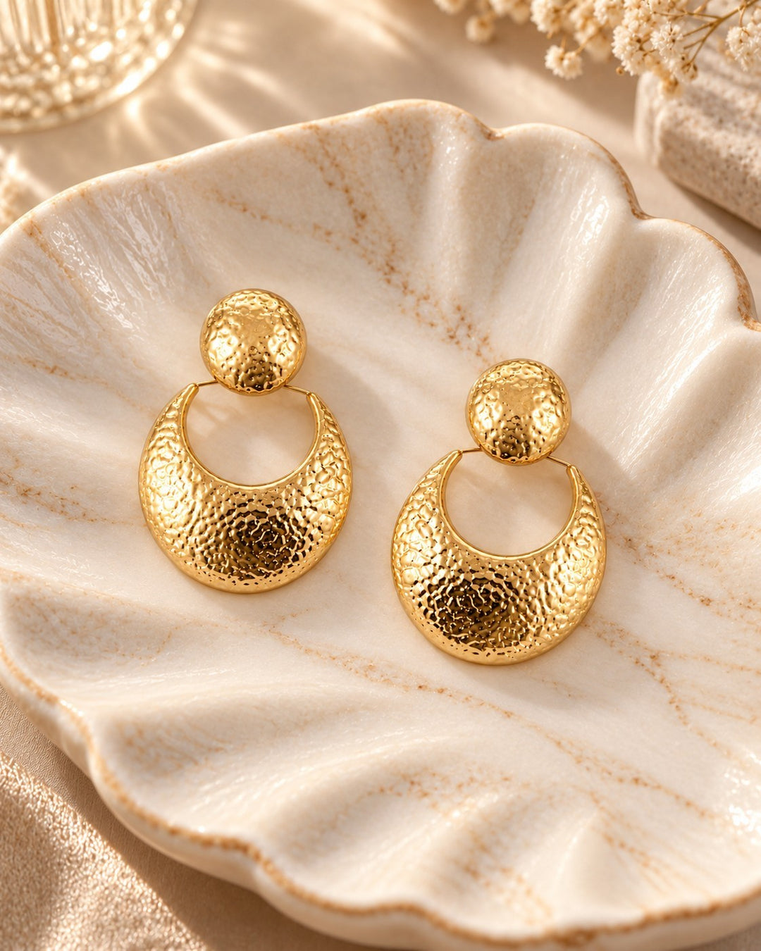 Elli Jewellery hammered 18ct gold plated crescent drop earrings with circular studs resting on a decorative shell dish.